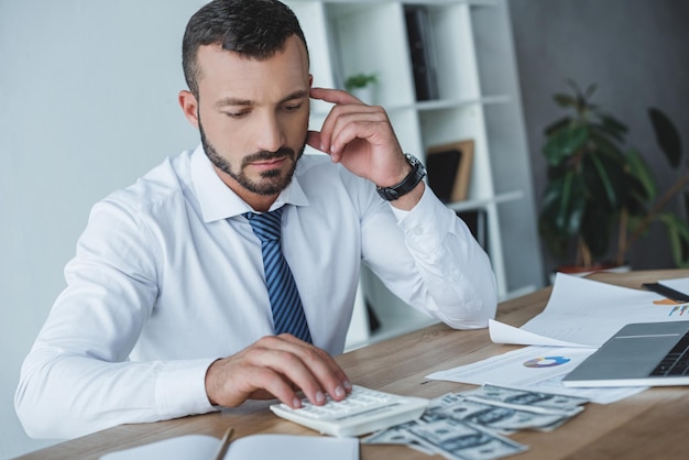 Pensive business adviser counting money with calculator in office