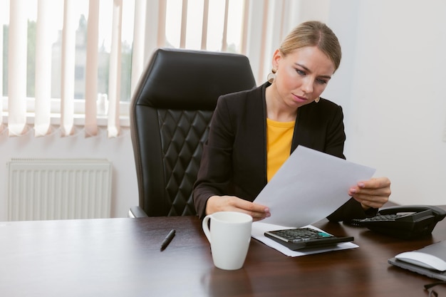 Pensive business lady boss looks attentively at a sheet of paper while sitting at a table in his office