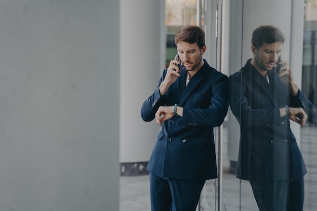 Pensive businessman in suit talking on mobile phone standing outside looking at his hand watch