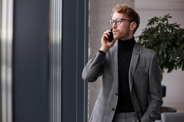 Pensive businessman wearing eyeglasses and stylish suit talking on mobile phone looking at window