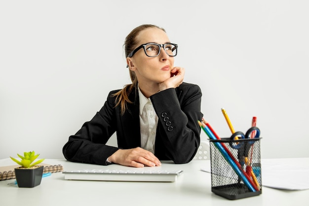 Задумчивая деловая женщина в куртке в очках с клавиатурой сидит на рабочем месте