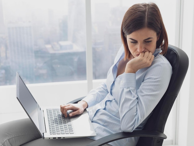Задумчивая деловая женщина, работающая в офисе с ноутбуком на коленях, она думает и планирует решение для своего бизнеса