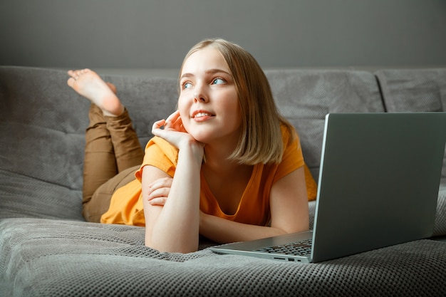 Pensive caucasian beautiful woman lies on sofa with laptop portrait of dreamy teenager girl thinking behind laptop at home in living room with crossed legs