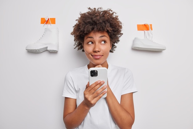 Pensive curly haired young woman holds chin considers something while using smartphone concentrated away wears casual t shirt poses against white backgroud with plastered shoes. Let me think