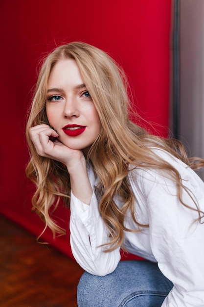 Pensive curly woman in shirt propping face with hand Charming girl in jeans sitting on red background