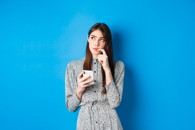Задумчивая милая девушка думает, как ответить на сообщение, задумчиво глядя в сторону и держа смартфон, стоя в платье на синем.