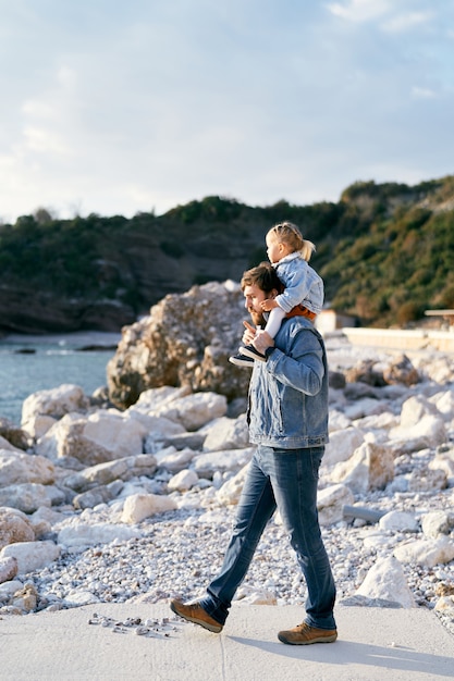 Pensive dad carries a little girl on his shoulders while walking along the pebble beach