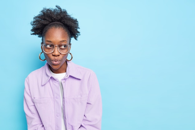 Pensive dark skinned curly millennial girl keeps lips rounded concentrated away being deep in thoughts 