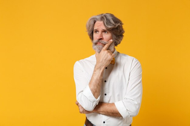 Pensive displeased elderly gray-haired mustache bearded man in white shirt bow tie posing isolated on yellow orange background. People lifestyle concept. Mock up copy space. Put hand prop up on chin.