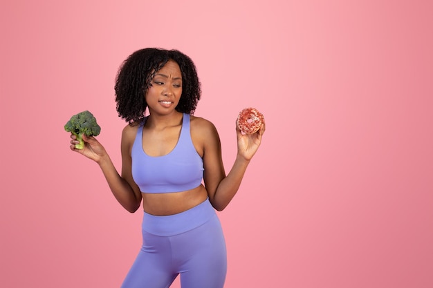 Pensive displeased hungry young african american lady in sportswear with broccoli looks at donut