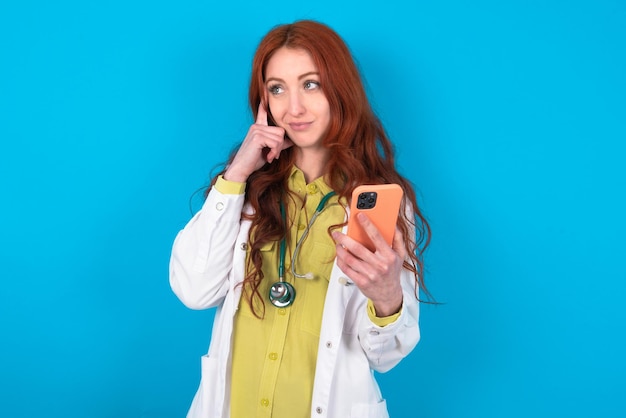 Pensive doctor woman keeps index finger on temple holds modern smartphone and concentrated aside