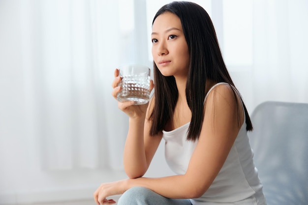 Pensive dreaming young asian cute woman think about bad\
relationships hold glass of water sit on chair in light office\
interior breakup from work relaxing time concept copy space\
offer