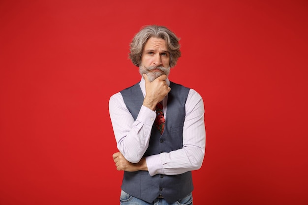 Pensive elderly gray-haired mustache bearded man in classic shirt vest colorful tie isolated on red background, studio portrait. People lifestyle concept. Mock up copy space. Put hand prop up on chin.