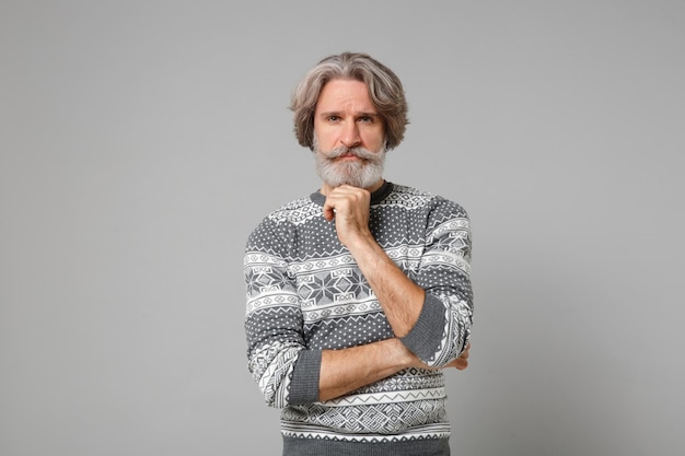 Pensive elderly gray-haired mustache bearded man in knitted\
sweater posing isolated on grey wall background, studio portrait.\
people lifestyle concept. mock up copy space. put hand prop up on\
chin.