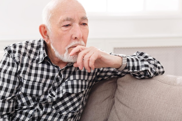 Pensive elderly man sitting on couch with thoughtful facial expression, copy space