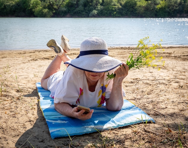 Pensive elderly woman, elderly pensioner, rest on the sand by the river