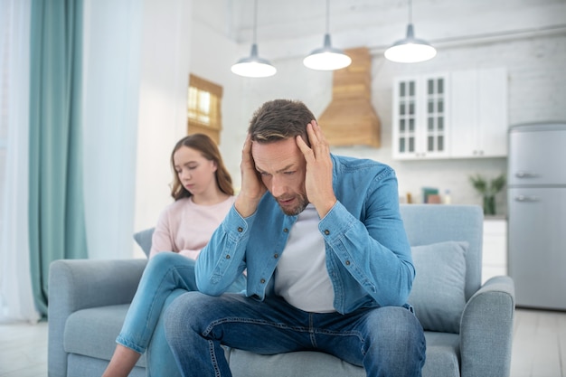 Pensive father clutching his head with his hands and a sad daughter sitting on the sofa at home.