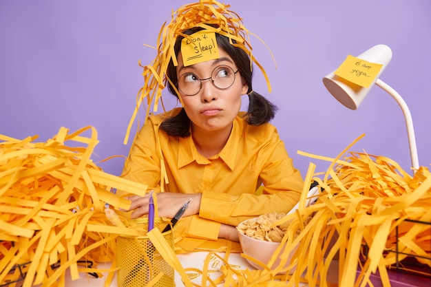 Photo pensive female office worker works on creative project thinks about finishing task has mess on desktop tries not to sleep to finish work in time isolated over purple wall