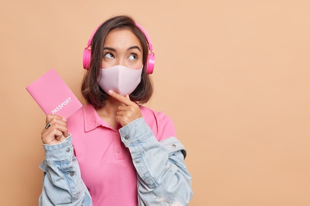 Photo pensive female traveler going to have fright abroad during coronavirus pandemic wears protective face mask holds passport listens music via headphones looks away wears t shirt and denim jacket