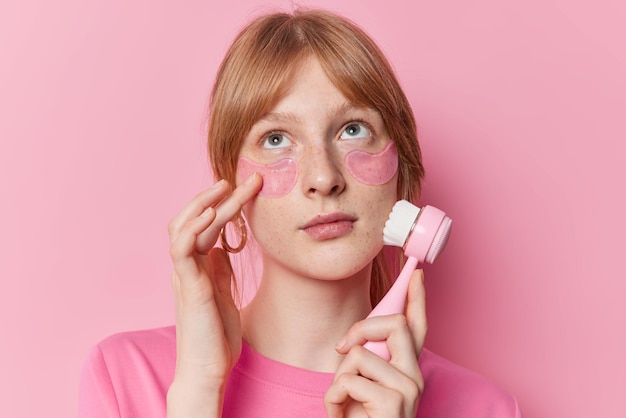 Pensive freckled millennial girl makes face massage with brush applies hydrogel patches under eyes concentrated above has ginger hair poses against pink background Facial treatments concept