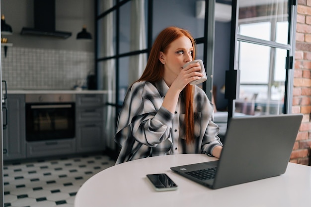 생각에 잠긴 프리랜서 여성은 원격 작업을 하거나 멀리 내다보는 테이블에 앉아 노트북 컴퓨터에서 공부하는 동안 커피 한 잔을 즐기고 있습니다.