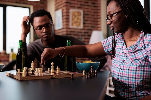 Pensive friends sitting at table in living room while playing chess together. Smart people relaxing at home enjoying fun strategy boardgames together while having snacks and beverages.