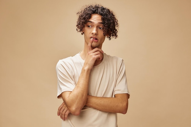 Pensive funny awesome tanned curly man in basic tshirt can not\
make decision posing isolated on beige pastel background fashion\
new collection offer people and emotions concept free place for\
ad