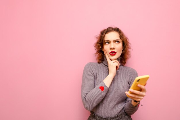 pensive girl in gray sweater stands on pink background with smartphone in hand and looks away