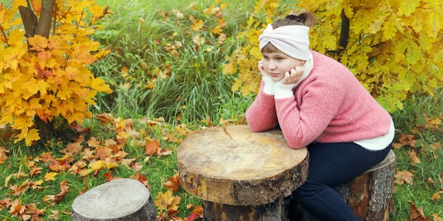 Pensive girl in pink sweater sits on wooden stump near autumn trees with yellow foliage