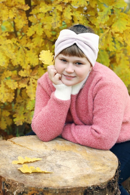 Pensive girl in pink sweater sits on wooden stump near autumn trees with yellow foliage