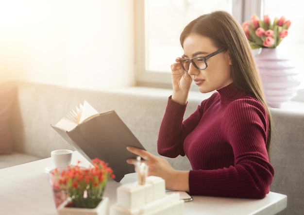 物思いにふける女の子がカフェのテーブルで本を読んでいます。休んでいる若い女性