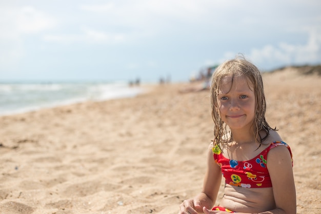 A pensive girl with wet blond hair in a swimsuit sits on the sandy seashore. Summer vacation, travel and tourism.