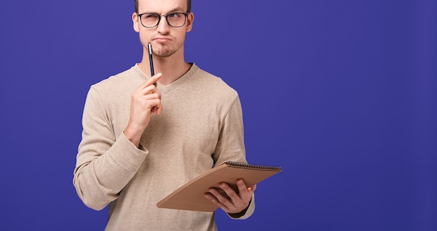 Pensive guy editor with brown notebook and black pen near his face looks straight