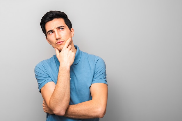 Pensive handsome man in blue shirt with hand on his chin looking up isolated against grey background with copy space