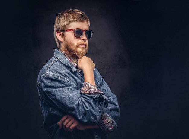 Pensive hipster guy wearing sunglasses dressed in jeans jacket posing with hand on chin. Isolated on a dark background.