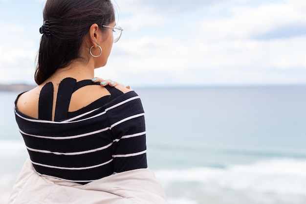 Pensive Hispanic woman touching painful shoulder on seashore