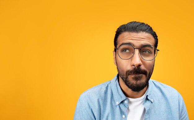 Pensive indian millennial man in glasses look at free space think isolated on orange background