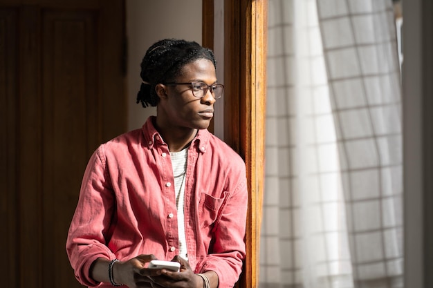 Pensive introverted African American man with mobile phone looking out window standing in house