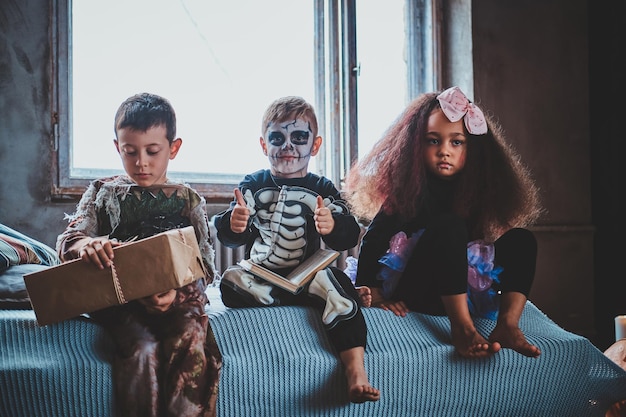 Pensive kids are sitting on the bed wearing Halloween costumes, boy is holding a box.