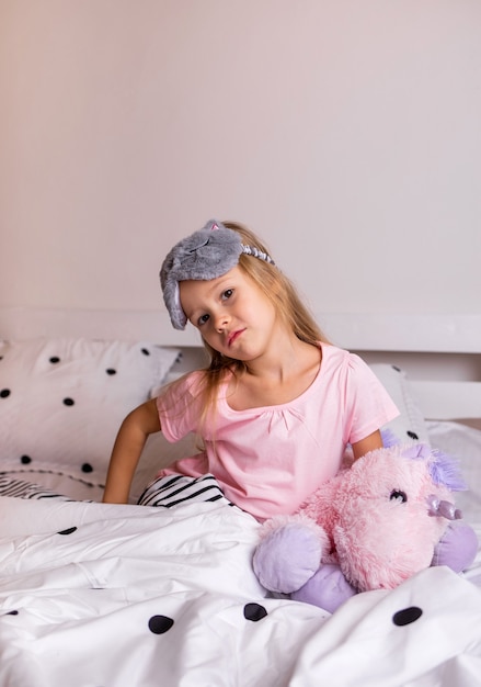 A pensive little blonde girl in pajamas is sitting on the bed linen with a soft toy on the bed in the room