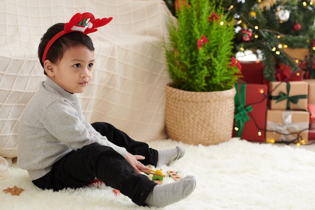 Pensive little boy wearing reindeer antlers headband sitting on the floor next to Christmas tree