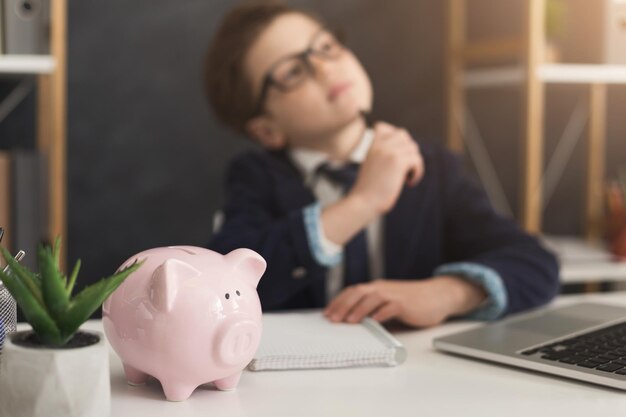 Pensive little businessman thinking of income at office table with piggy bank, selective focus