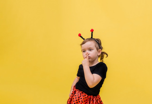 pensive little girl in a Ladybird costume points her finger "Quietly" against a yellow isolated