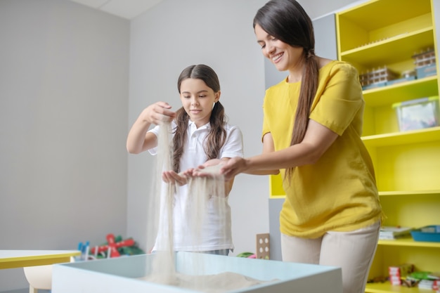 Pensive long-haired school-age girl with cute smiling woman in sand therapy session