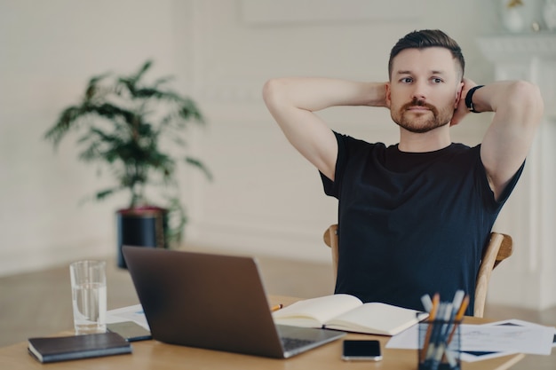 Pensive male freelancer takes break keeps hands behind head poses at desktop works on laptop computer has thoughtful expression thinks about future plans plans week dressed in black t shirt.