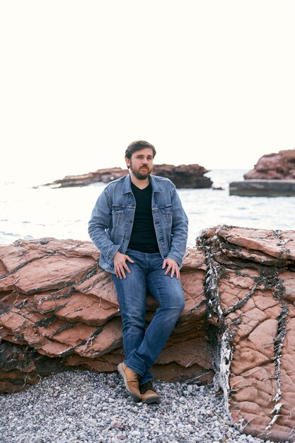 Pensive man in jeans and denim jacket posing standing on the shore leaning on stones against the