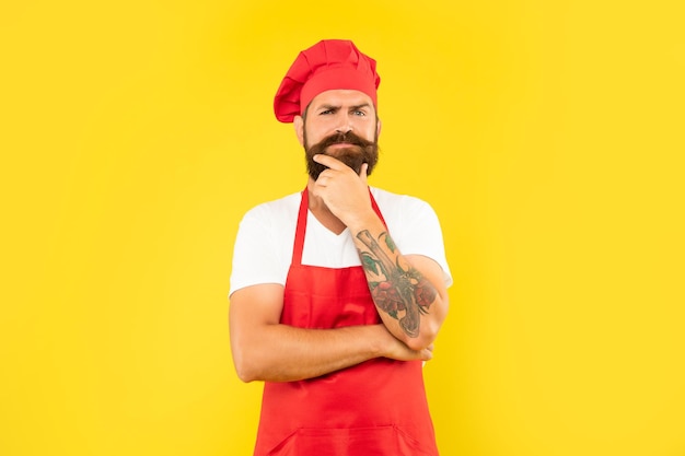 Pensive man in red toque and apron stroking chin yellow background chef