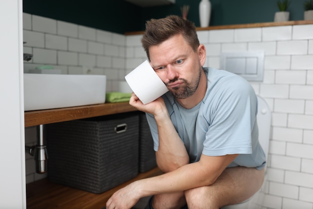 Pensive man sits on toilet with a roll of toilet paper.