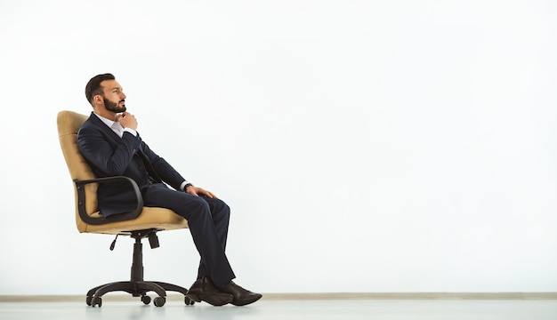 The pensive man sitting on an office chair on the white wall background