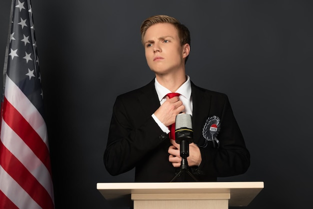 Pensive man touching tie on tribune with american flag on black background